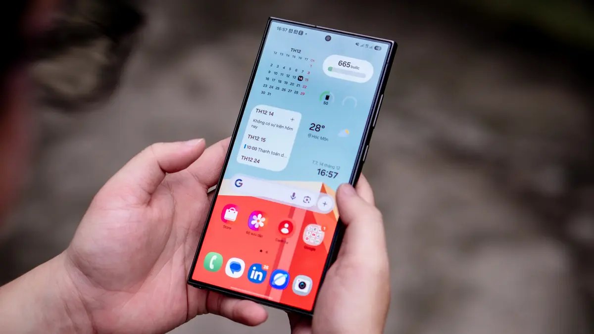 Hình ảnh một người đang cầm trên tay chiếc điện thoại Samsung Galaxy chạy giao diện One UI 7. Màn hình chính hiển thị các widget tiện ích như lịch, thời tiết, số bước chân, cùng với thanh tìm kiếm Google ở phía trên.